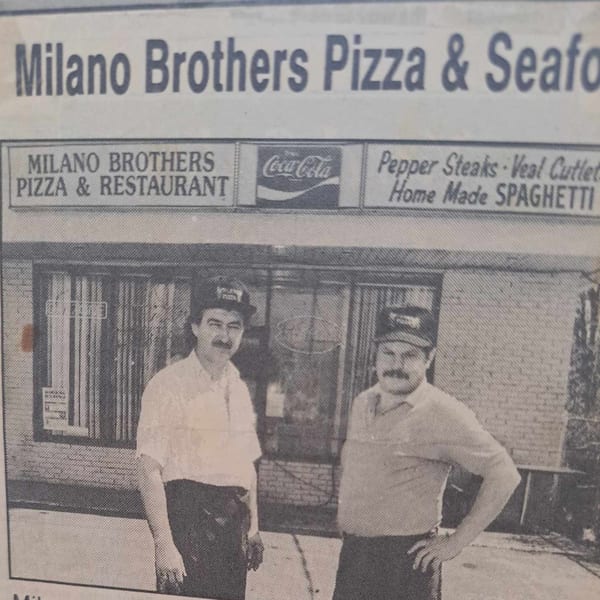 Two men in hats and aprons stand outside Milano Brothers Pizza & Restaurant, a brick building with signs.