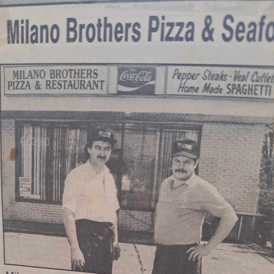 Two men in hats and aprons stand outside Milano Brothers Pizza & Restaurant, a brick building with signs.